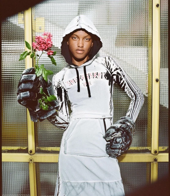 Fashion model wearing a hooded top and skirt, holding a rose and wearing large gloves. Studio lighting, against a paneled background.