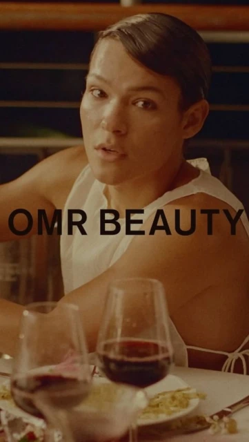 Close-up of a person seated at a table with wine glasses and dinnerware, against a background with the text 'OMR BEAUTY'.