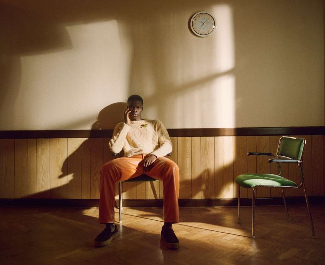 Young black man in a cream-colored sweater and orange pants sits indoors beneath a clock, next to a vintage green chair in warm lighting.