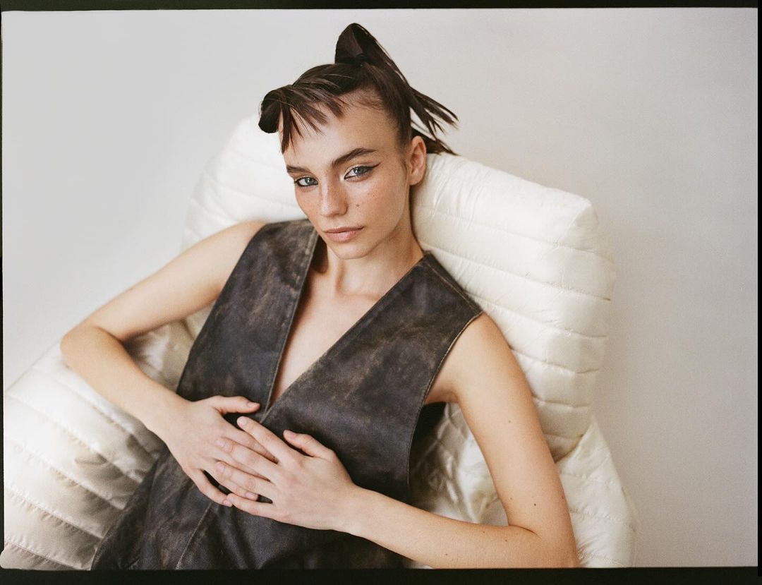 Stylish adult woman with unique hairstyle poses in a black vest against a white padded chair. The image evokes a blend of fashion and art.