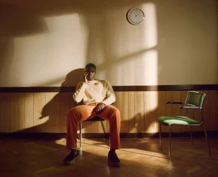 A Black man in a cream cable-knit sweater and orange trousers seated on a chair in a room with wood paneling and a wall clock.