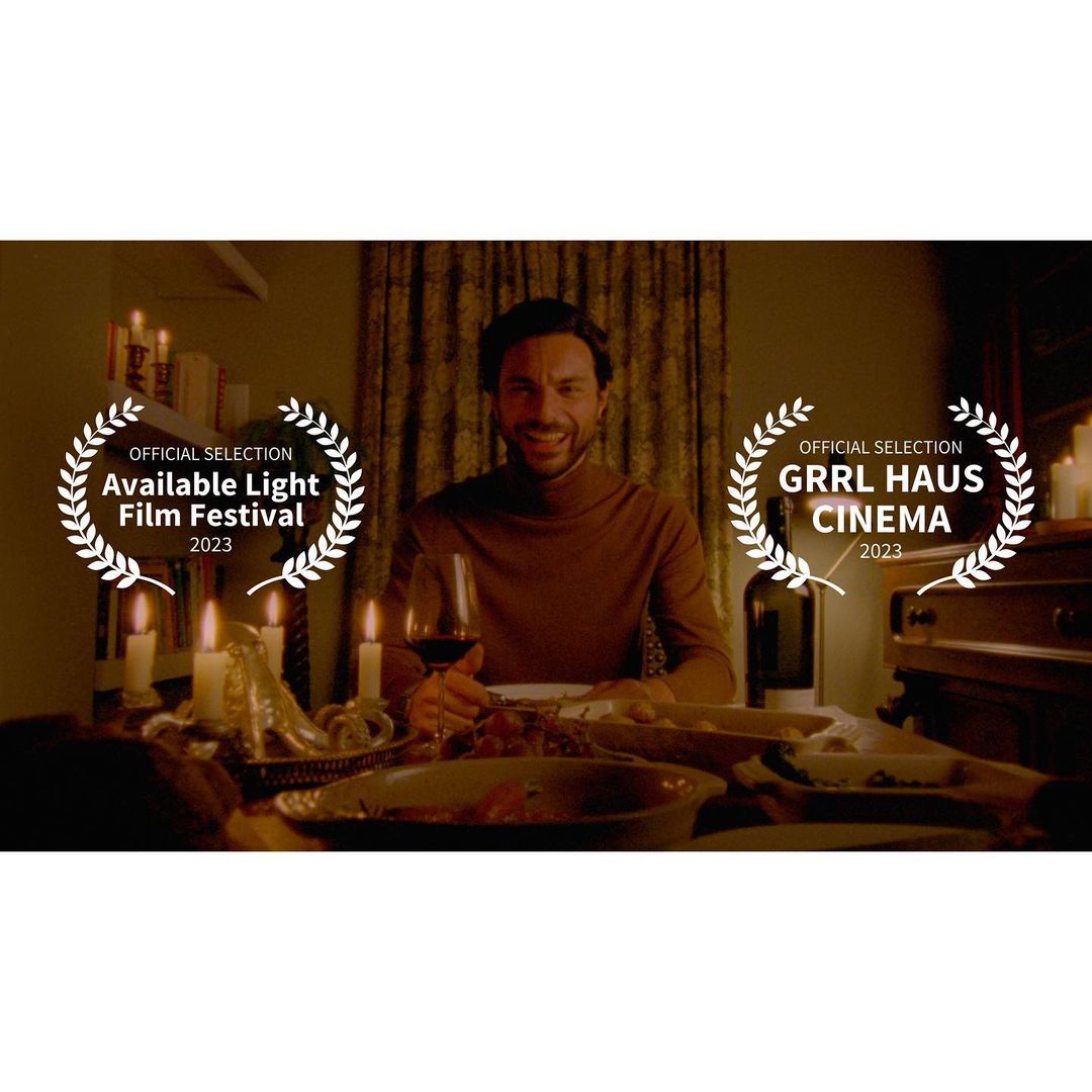 Man sits at a table with food and wine in a dimly lit room. Laurel wreath film festival logos are on either side of the man.
