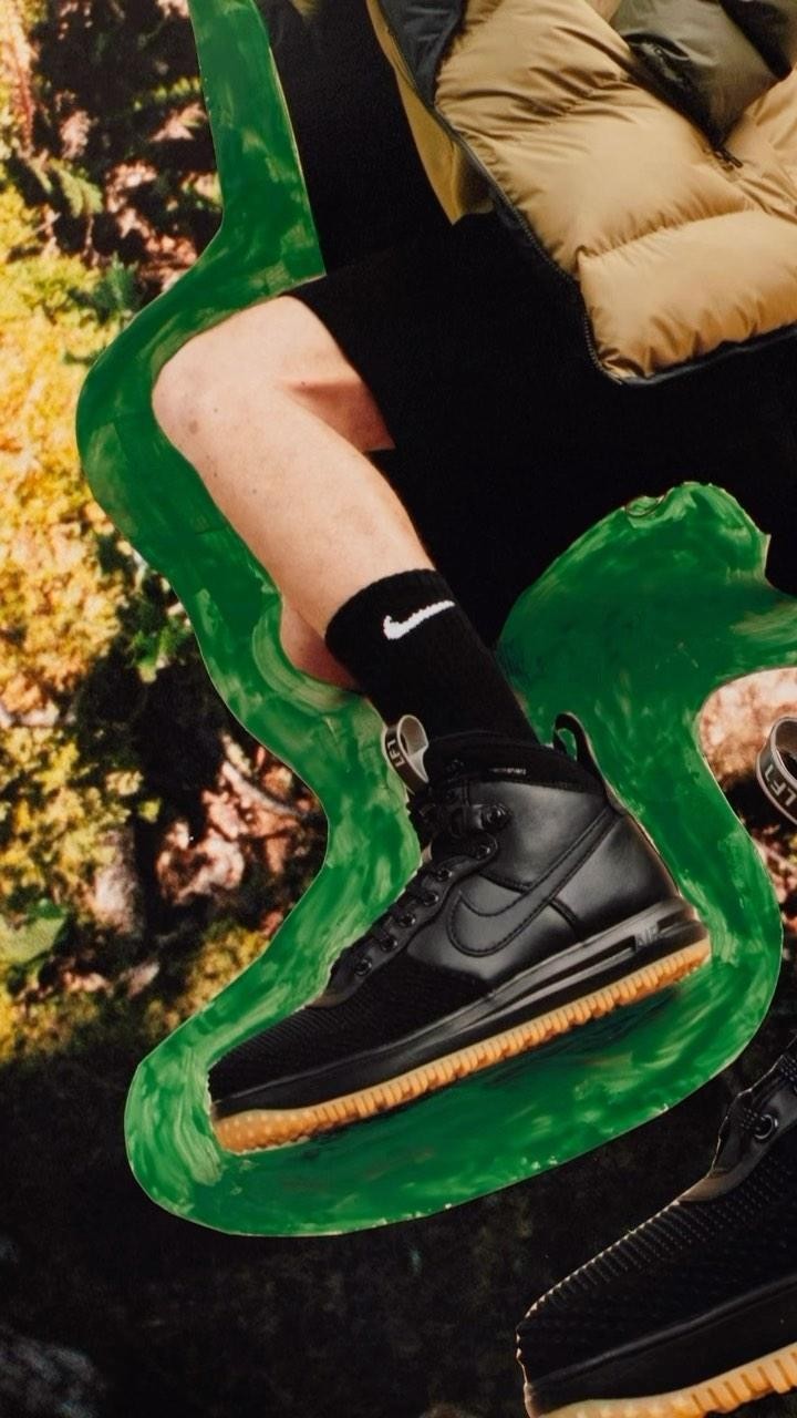 Black Nike boot with tan sole on a green surface, model leg with black Nike sock, green abstract paint streak, forest background.