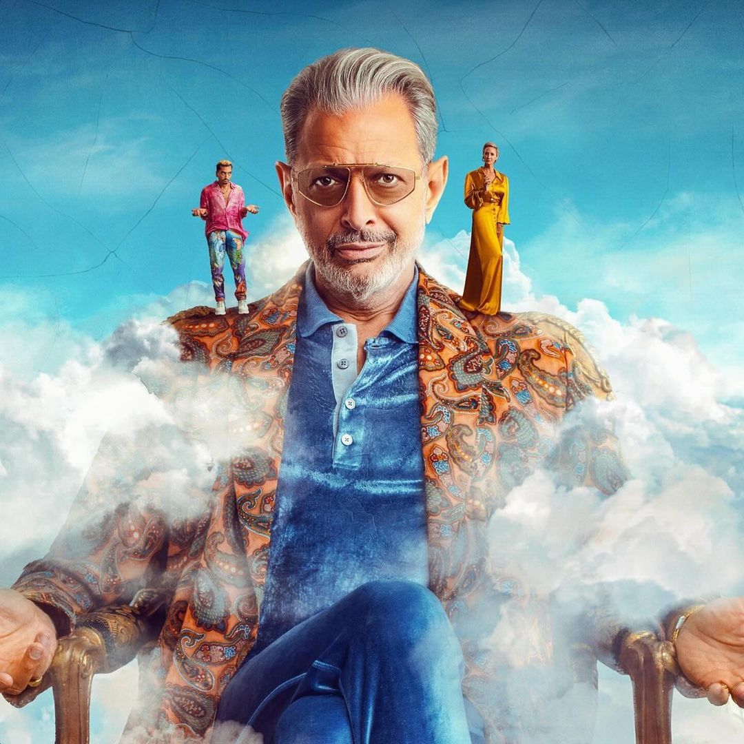 Portrait of Jeff Goldblum on a chair in a paisley blazer, with two people standing on his shoulders, against a cloudy sky backdrop.
