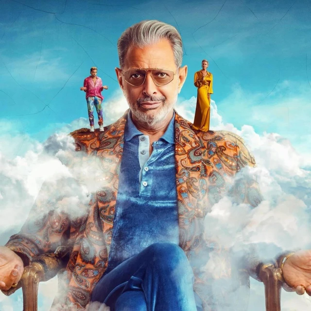 Portrait of Jeff Goldblum on a chair in a paisley blazer, with two people standing on his shoulders, against a cloudy sky backdrop.