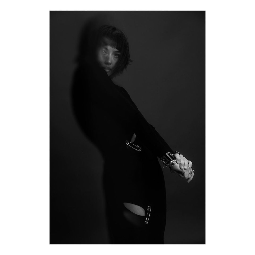 Monochrome studio portrait of a slender adult female model posing in a long black dress with safety pin accents on dark grey backdrop.