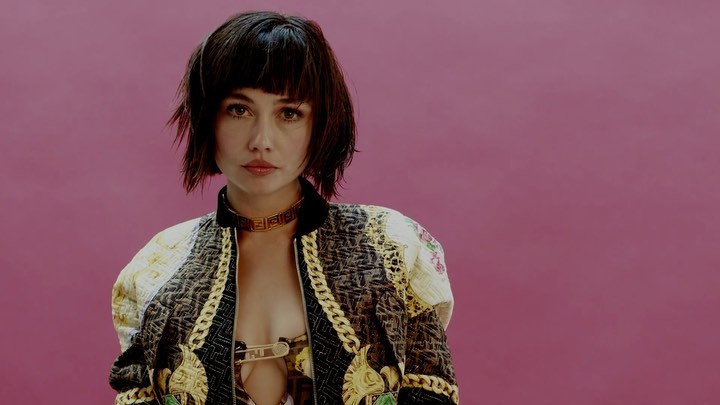 Portrait of a brunette woman with bangs wearing a decorative bomber jacket and collar against a mauve pink studio backdrop.