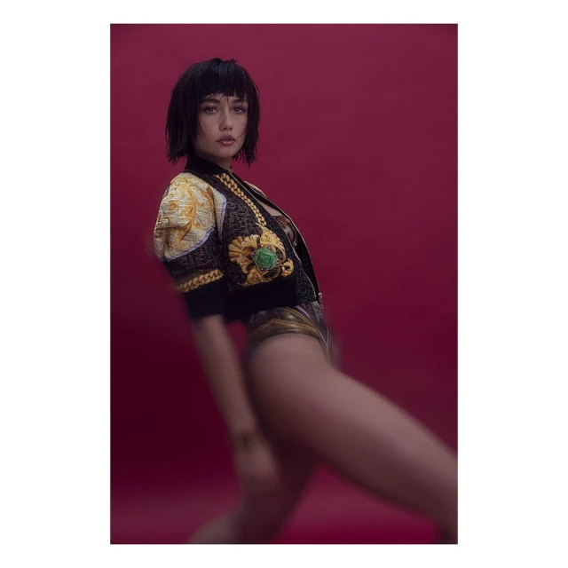 Brunette woman poses in a gold and black embellished jacket against a solid red background in a studio shot. Fashion photography.