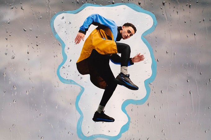 Athletic man in blue and yellow jacket leaps through a raindrop-covered window, showcasing dynamic energy and active lifestyle in motion.