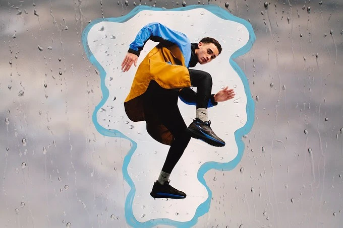 Athletic man in blue and yellow jacket leaps through a raindrop-covered window, showcasing dynamic energy and active lifestyle in motion.