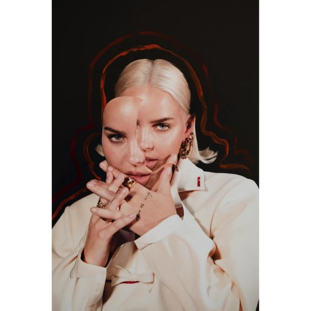 Portrait of a woman with light blonde hair and fair skin, wearing a white shirt and gold hoop earrings, set against a dark background.