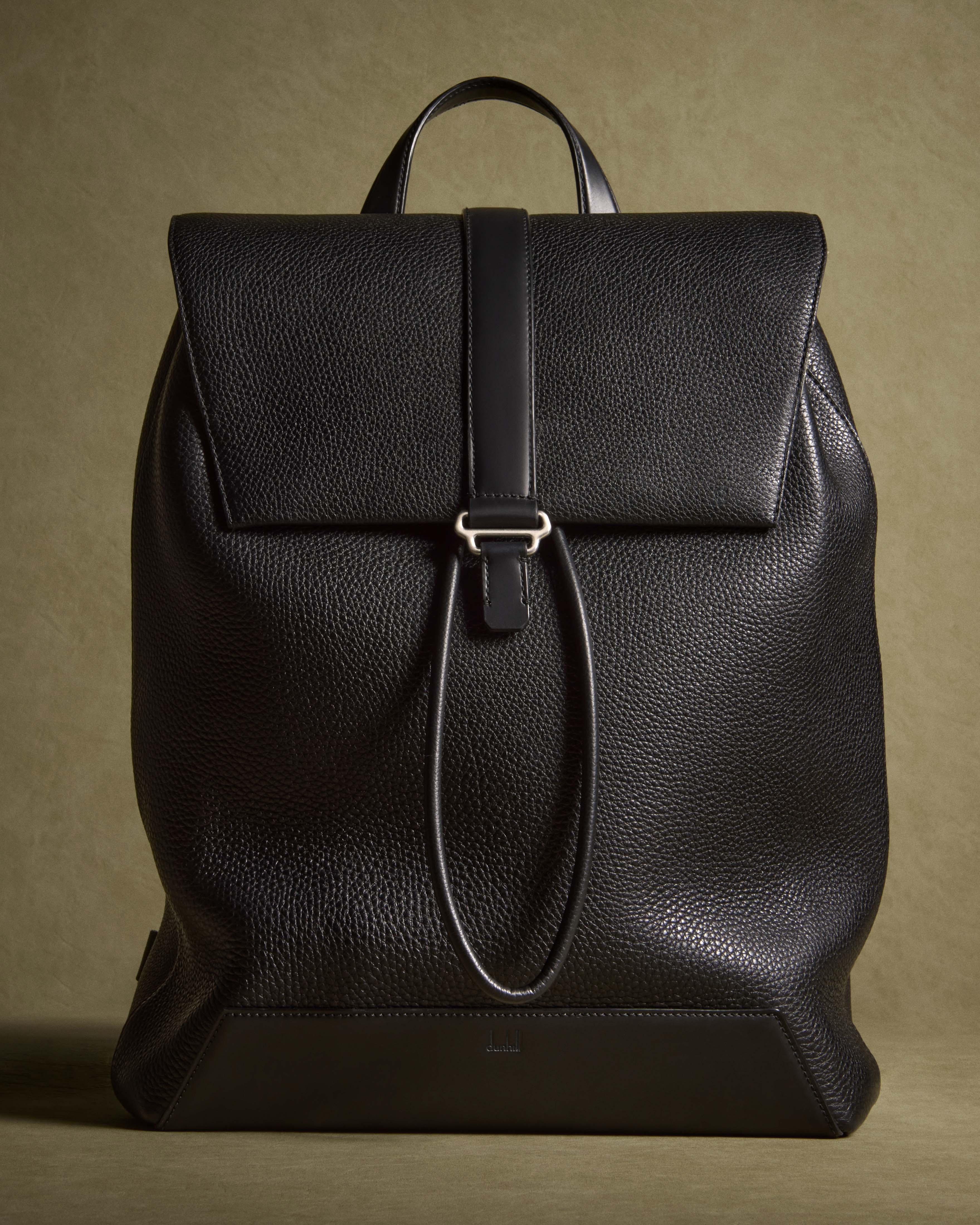 Studio shot of a black leather backpack with flap closure and drawstring, featuring silver hardware against an olive green backdrop.