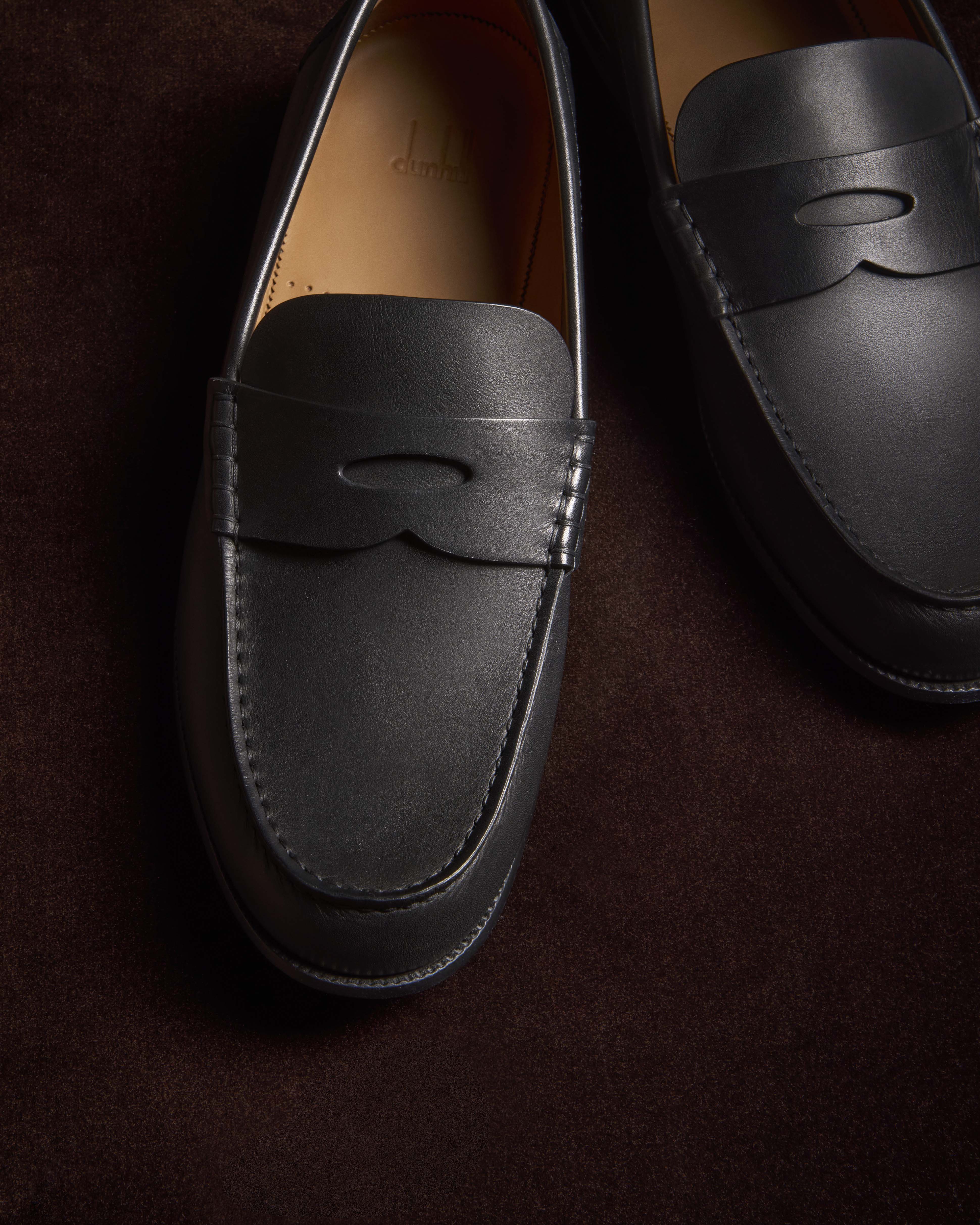 Elevated view of black leather penny loafers on a brown suede background, highlighting the formal men's footwear.