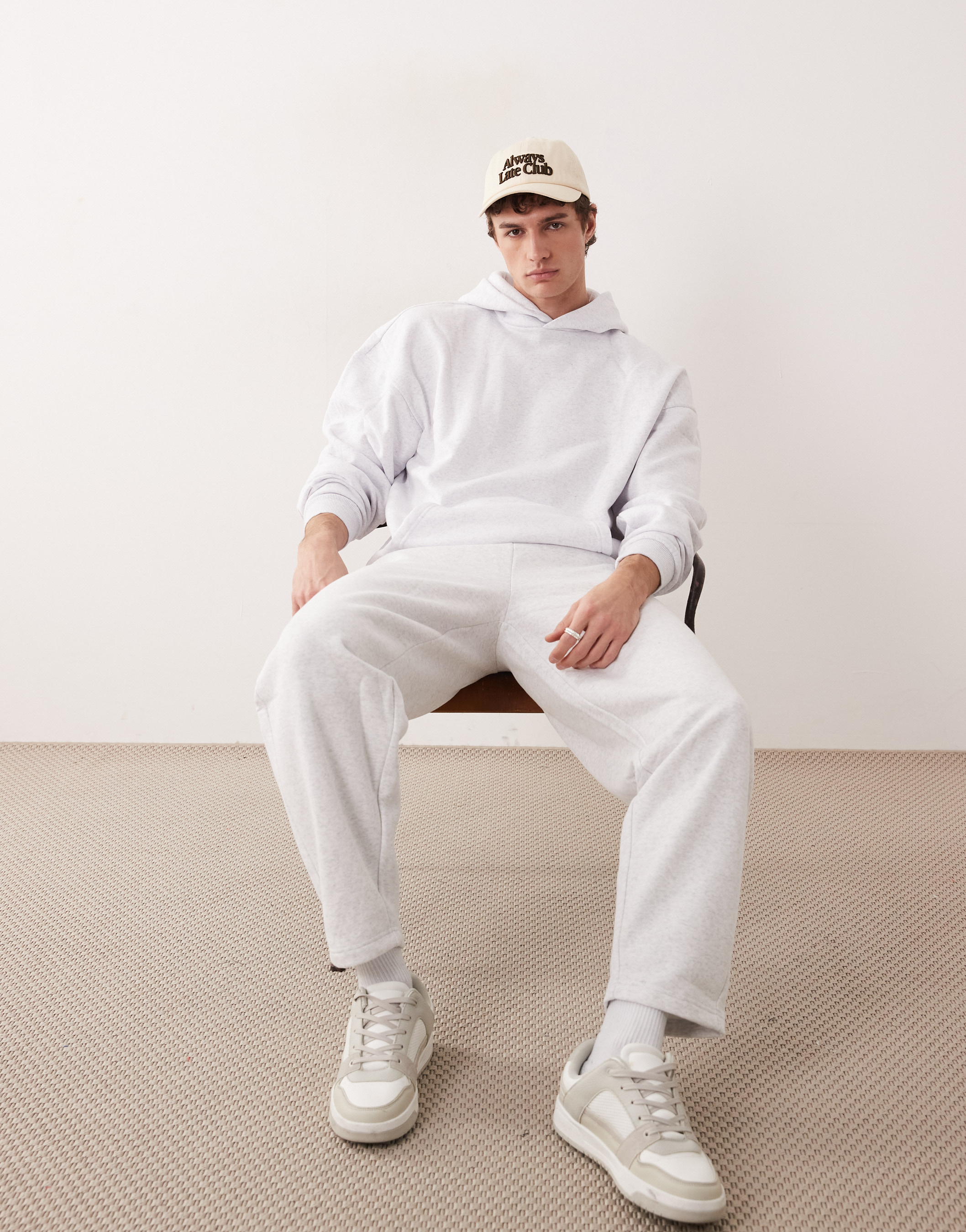 Young adult man in relaxed pose wearing matching grey hoodie and sweatpants, tan baseball cap, and sneakers sitting in a studio setting.