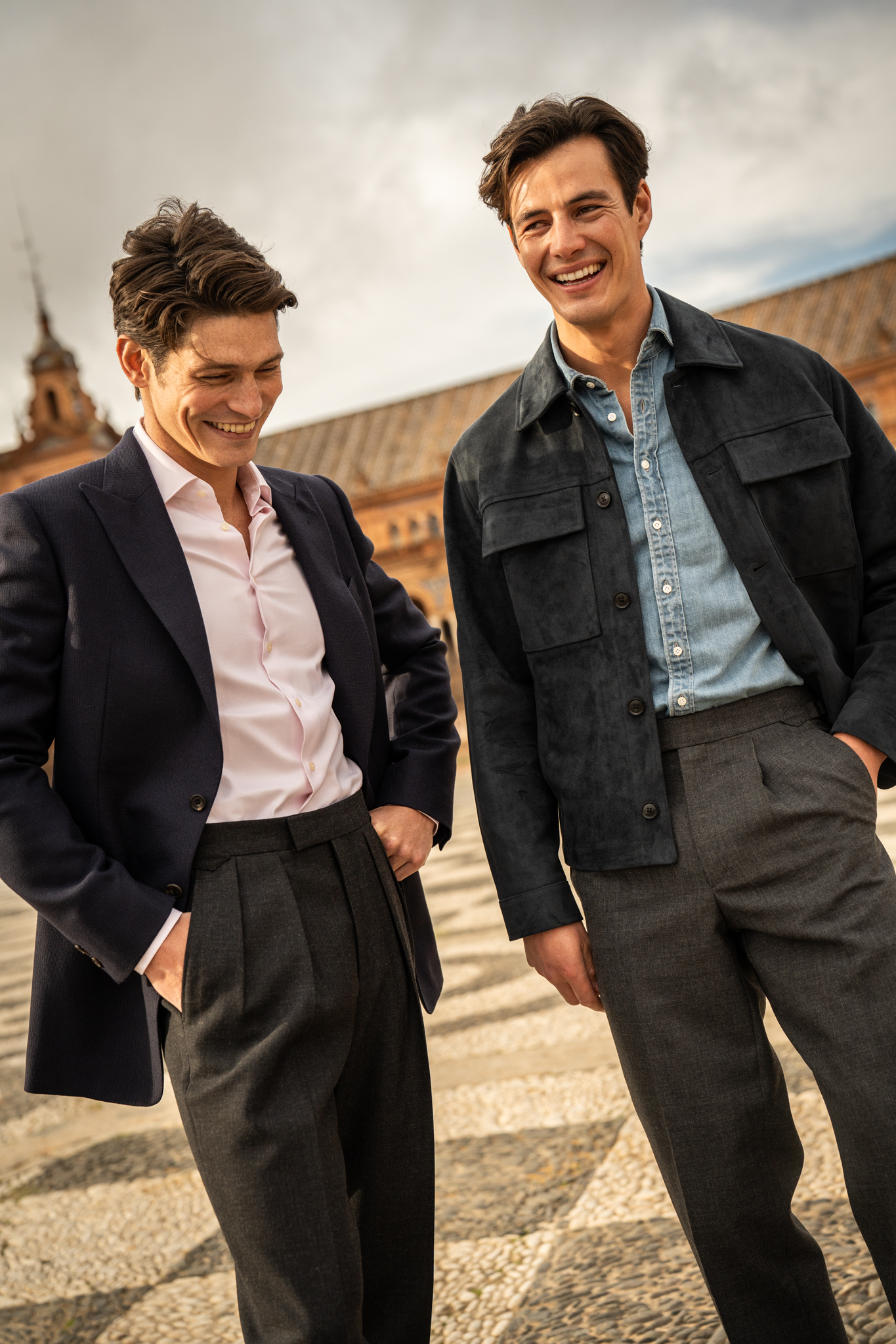 Two adult men in formalwear stand outdoors with warm lighting. One laughs while the other looks downward, with a plaza background.