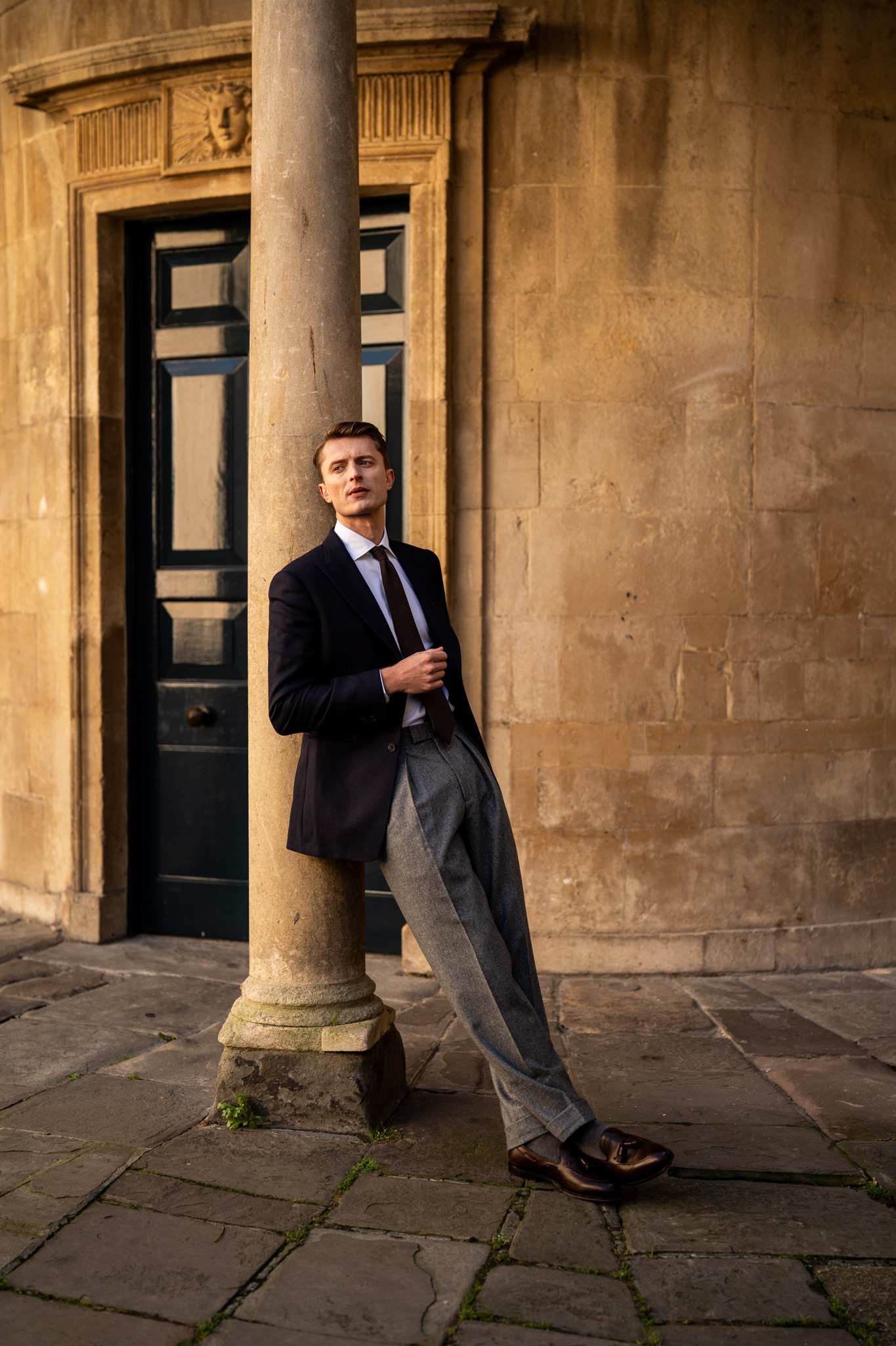 An adult male is posing leaning against a stone column in a blazer, dress pants, dress shoes and a tie, with a building facade behind him.