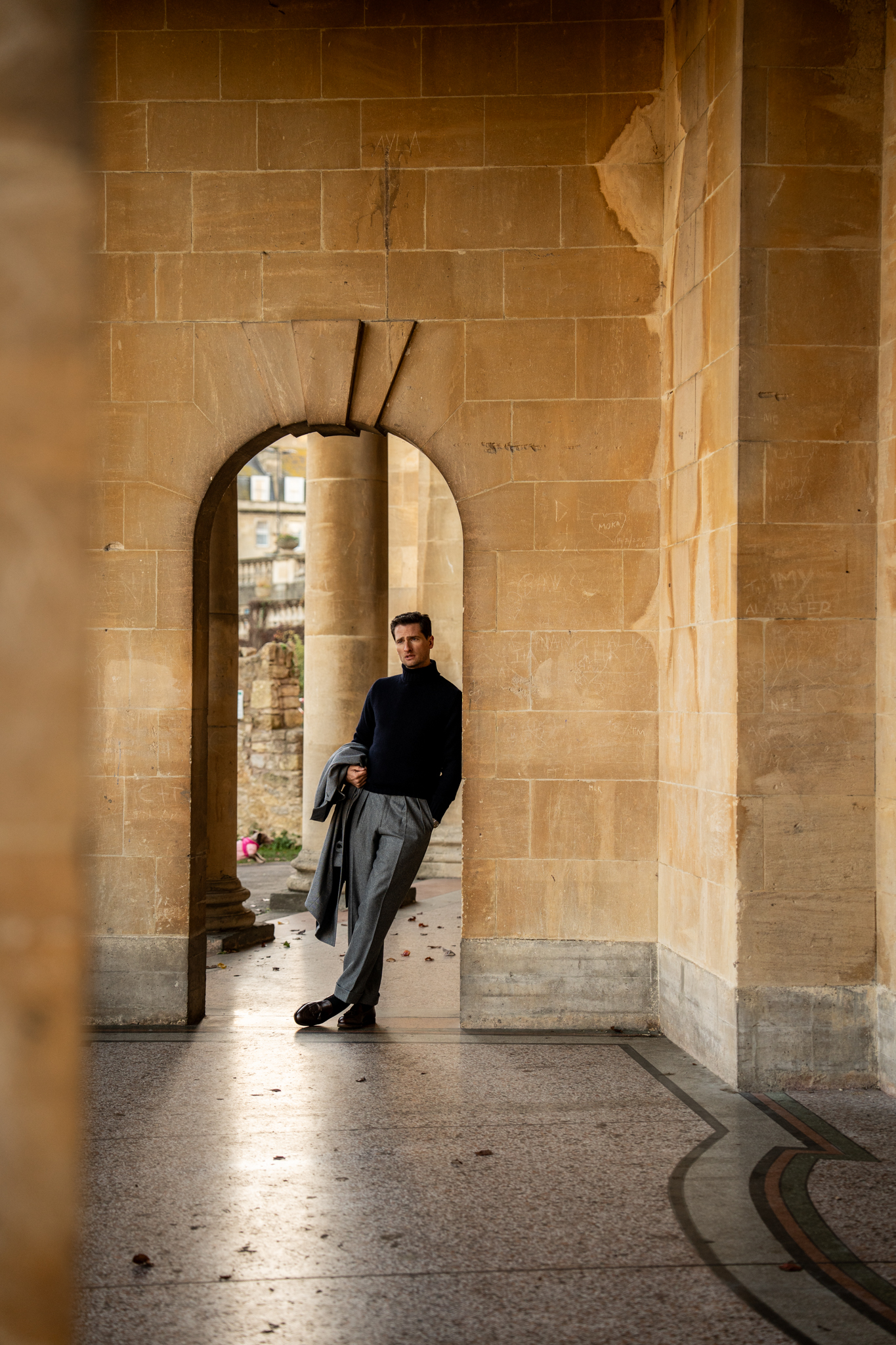Man in a black sweater and grey pants stands in a stone archway holding a jacket, with a partially visible city in the background.