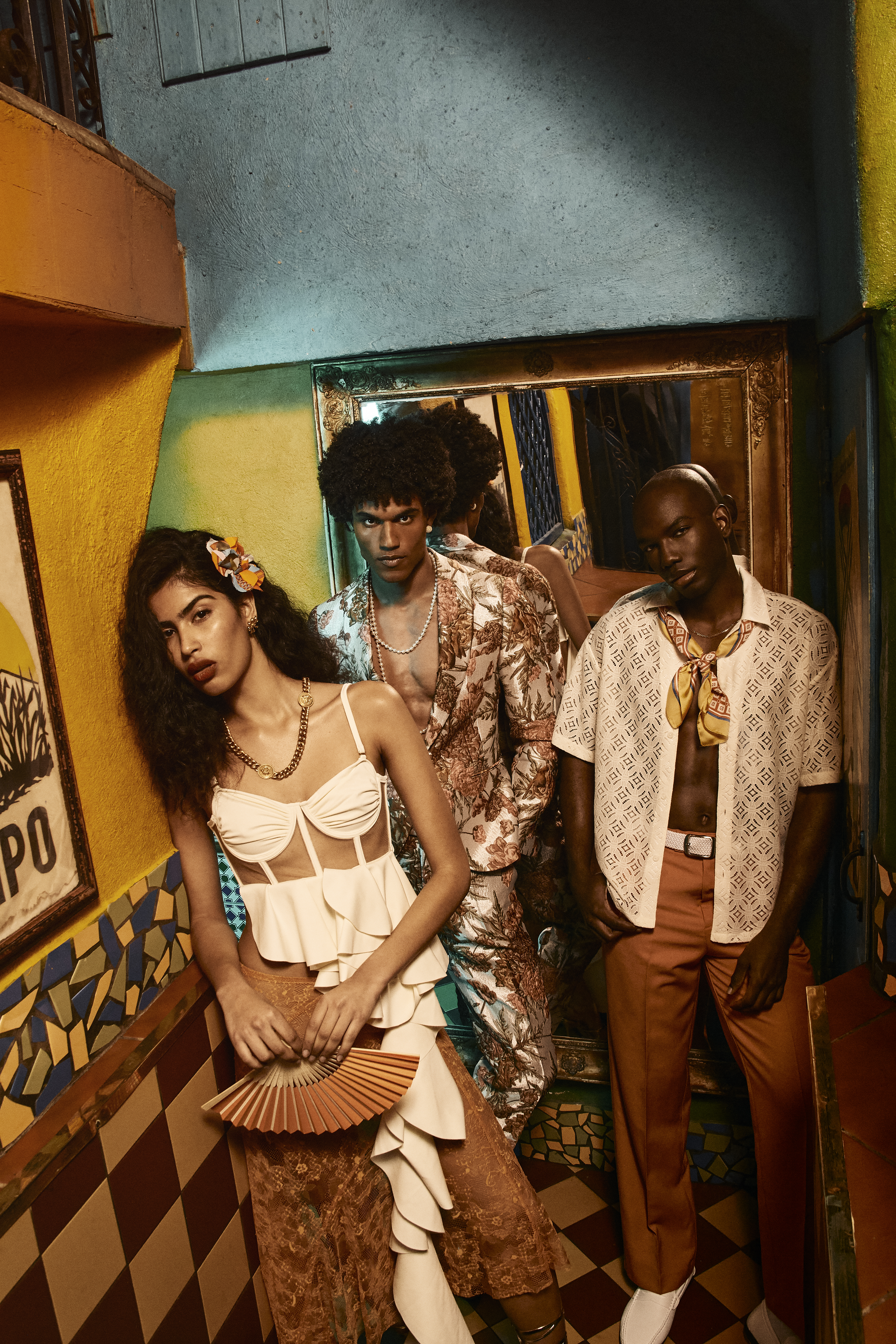 Three fashion models pose indoors; a woman in white holding a fan, surrounded by two men in patterned tops and earth-tone pants.