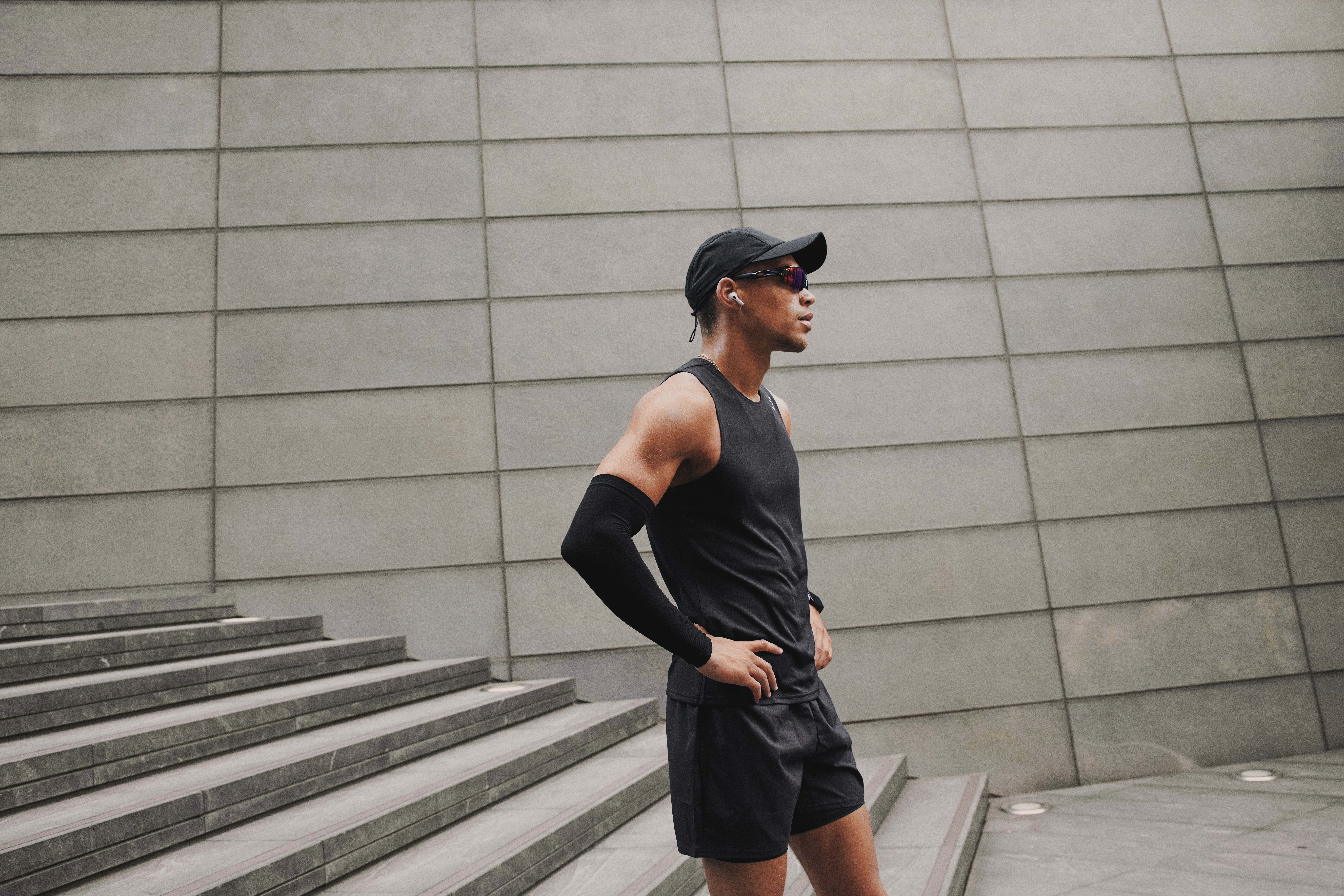 Athletic man in workout gear stands on stone steps against a gray wall, wearing a baseball cap and sunglasses in an urban setting.