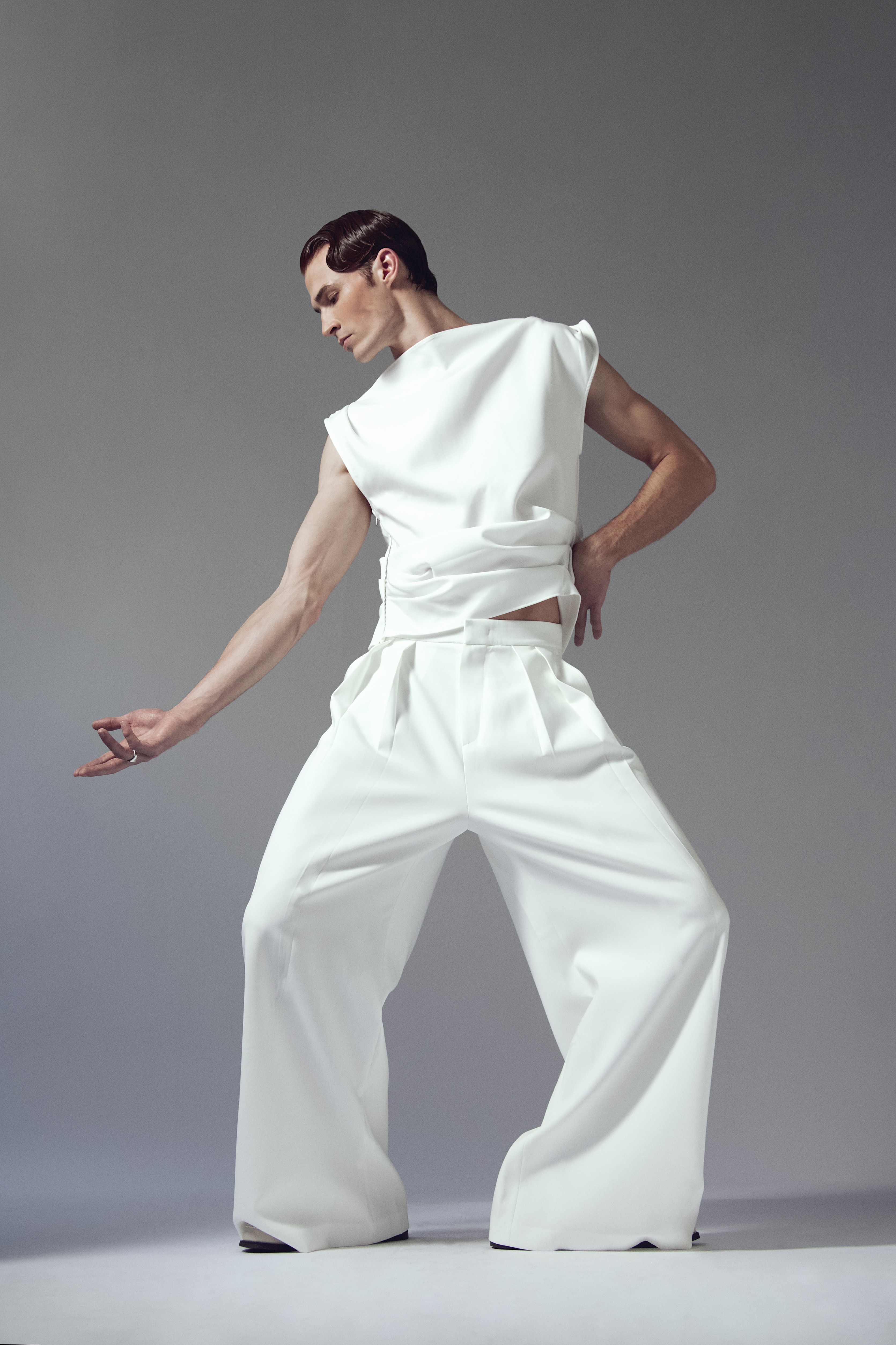 A slender adult male poses in a sleeveless white top and wide-leg white pants against a gray background in a fashion shot.