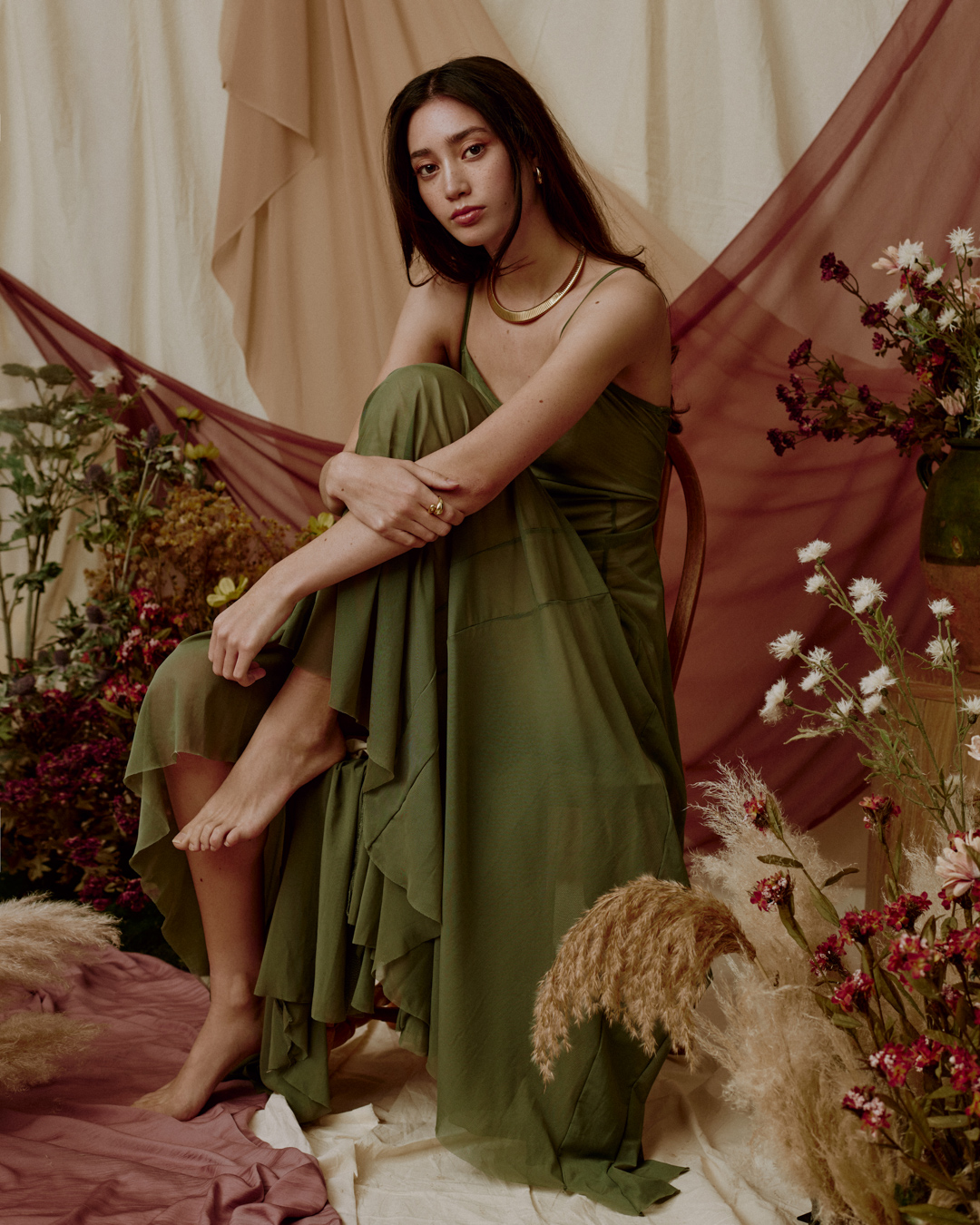 A long-haired woman in an olive-green dress sits barefoot amid flowers and draped fabrics, with a neutral studio backdrop and soft lighting.
