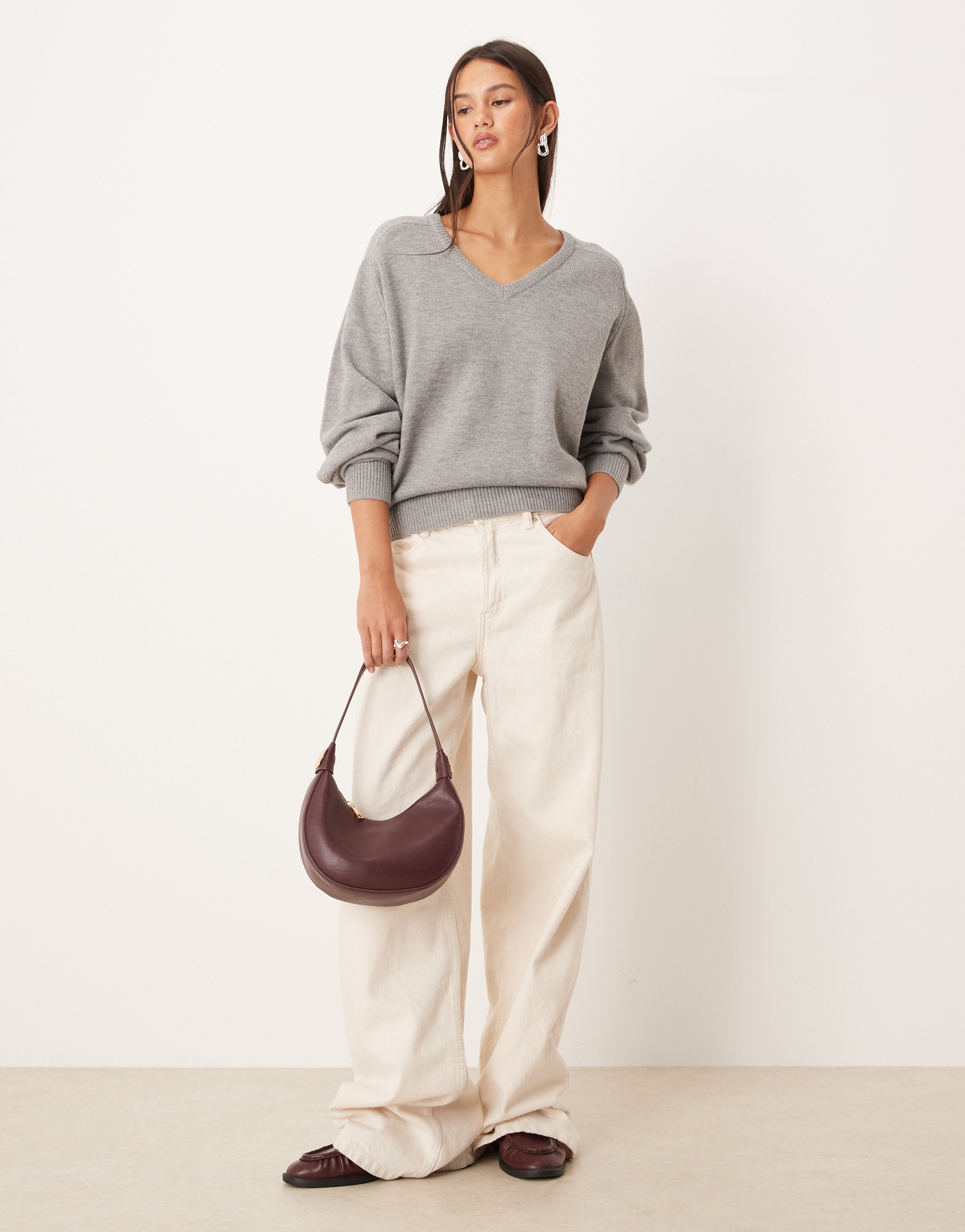 A woman models a gray sweater and white pants, holding a burgundy-colored bag in a minimalist studio setting, full-length.