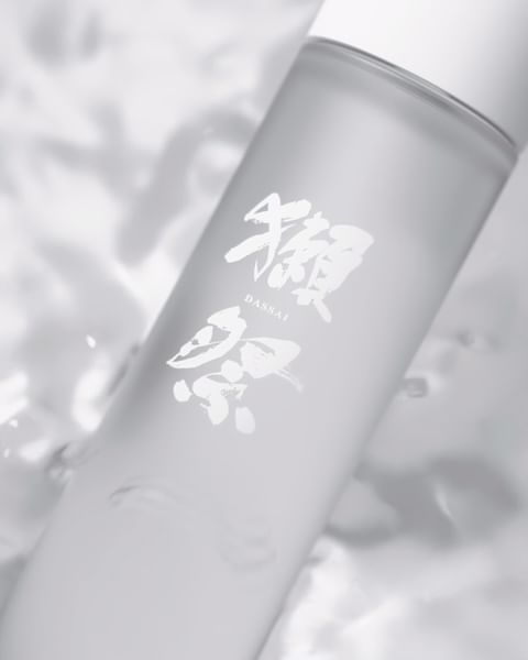 A bottle of Dassai sake is displayed against a blurred background with subtle textures and a minimalist aesthetic in a soft, diffused light.