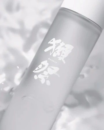 A bottle of Dassai sake is displayed against a blurred background with subtle textures and a minimalist aesthetic in a soft, diffused light.