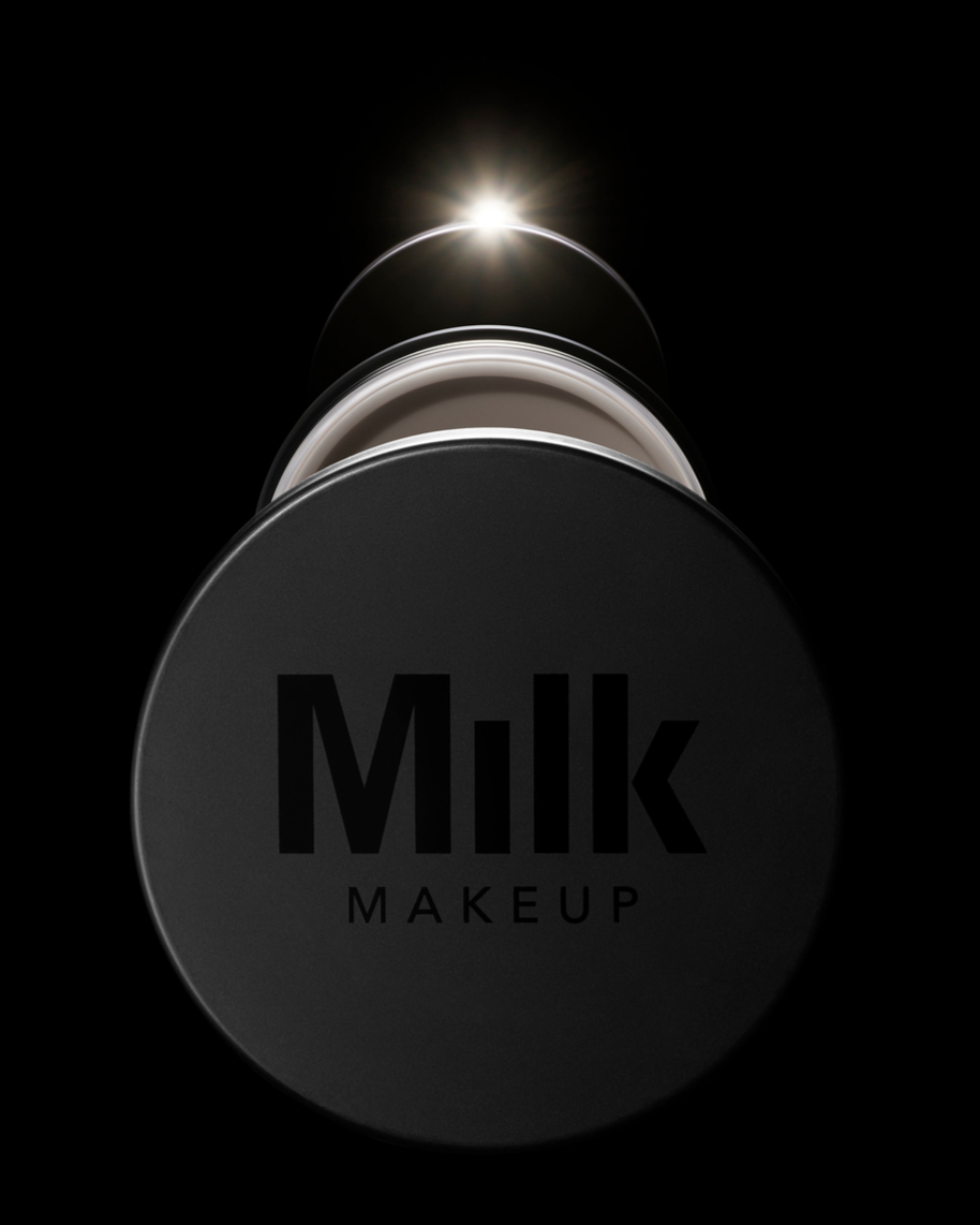 Close-up of Milk Makeup product container with the brand's logo on the lid against a stark black background and a bright light source.