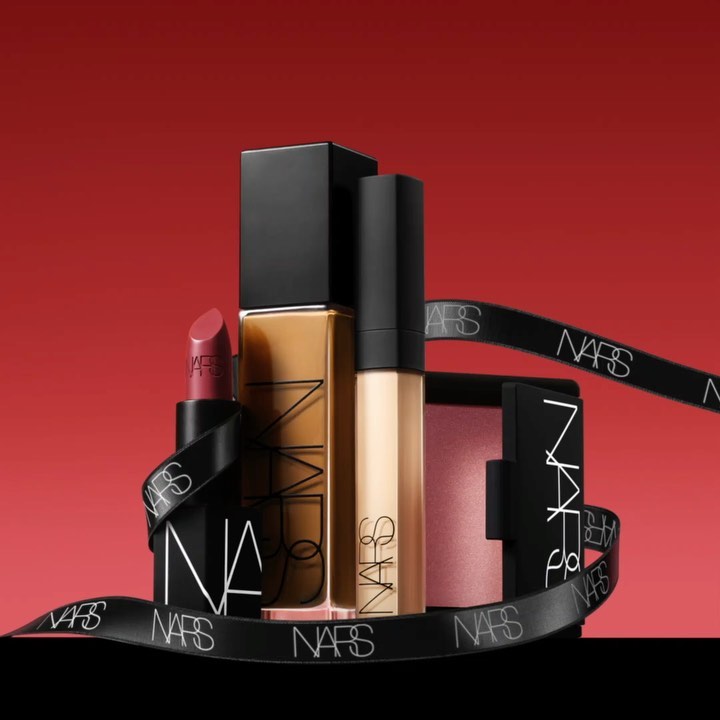 NARS Cosmetics makeup set featuring lipstick, foundation, concealer, blush, and a ribbon with the NARS logo, against a red backdrop.