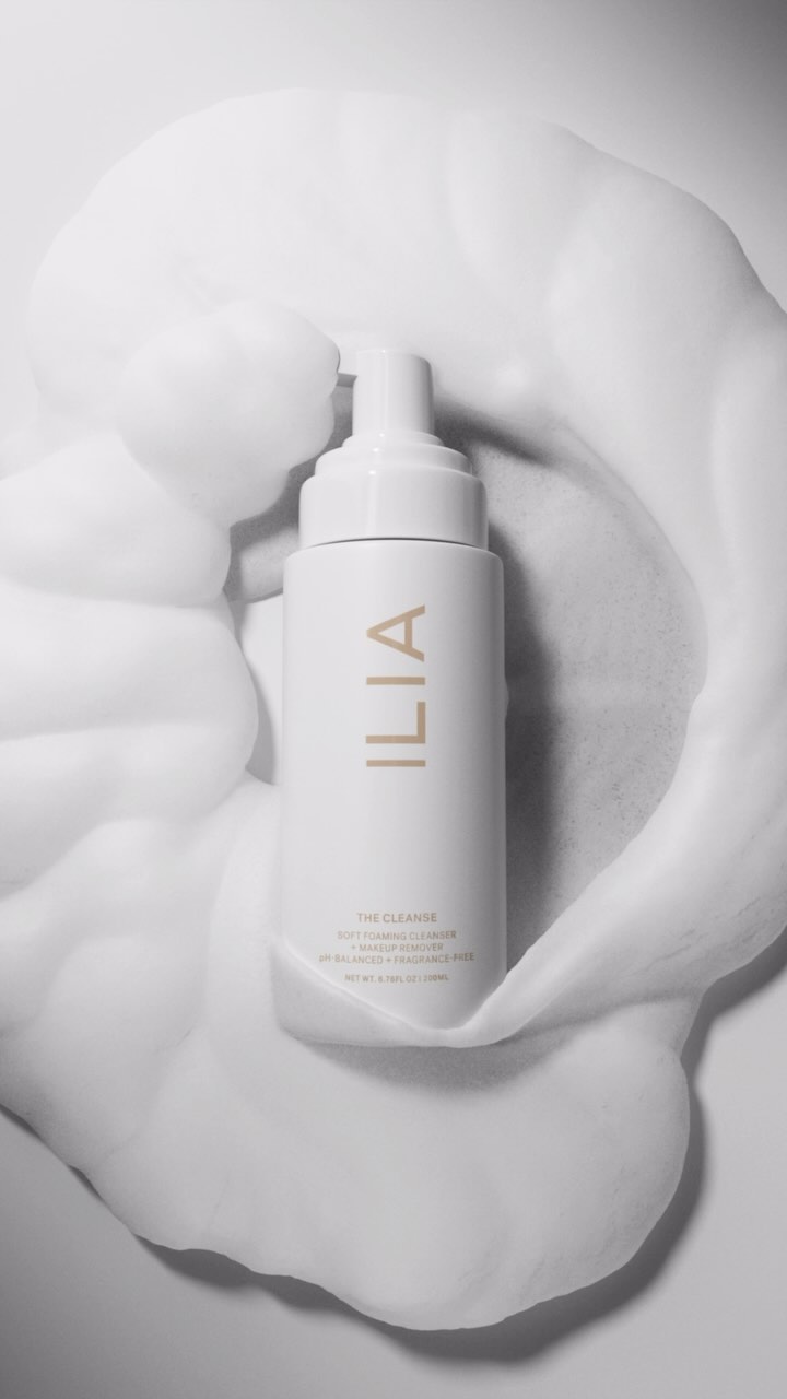 Elevated view of white Ilia cleanser bottle surrounded by white foam, set against a white backdrop, in a beauty product shot.