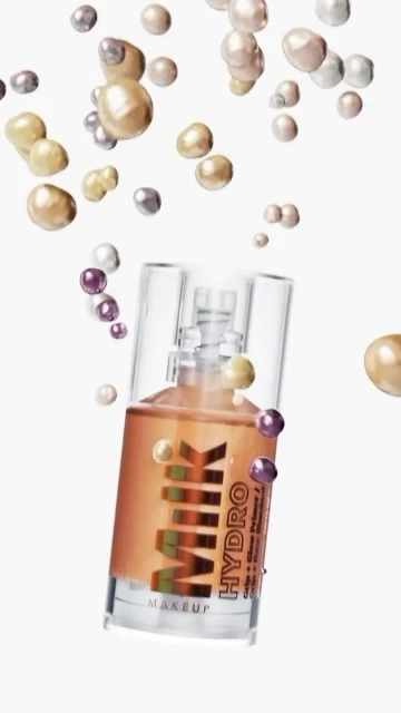 Close-up of a MILK Makeup Hydro Grip Primer bottle surrounded by floating iridescent pearls on a white background. Beauty and skincare.
