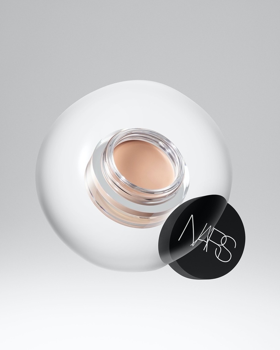 Close-up of NARS pot of beige concealer on a clear spherical surface, set against a gradient white backdrop in a beauty shot.