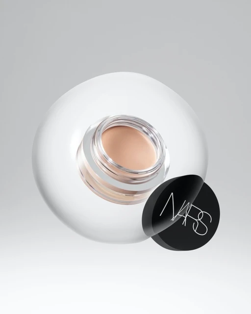 Close-up of NARS pot of beige concealer on a clear spherical surface, set against a gradient white backdrop in a beauty shot.