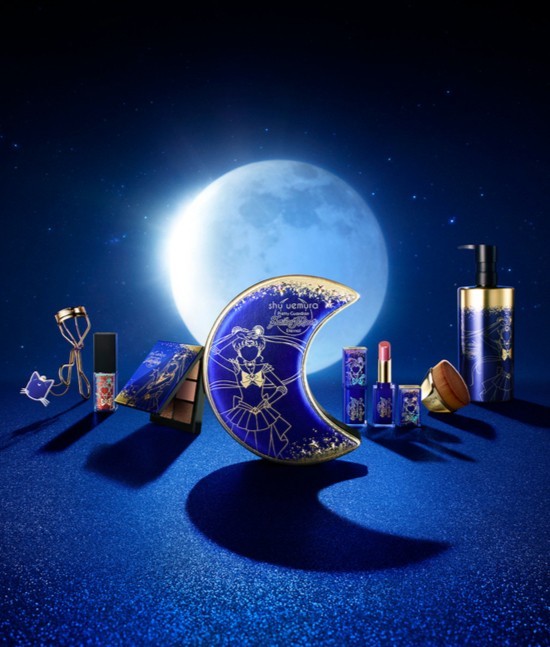 Shu Uemura x Sailor Moon makeup collection featuring eye shadow palette, lipstick, cleanser, and eyelash curler, presented against a blue backdrop.