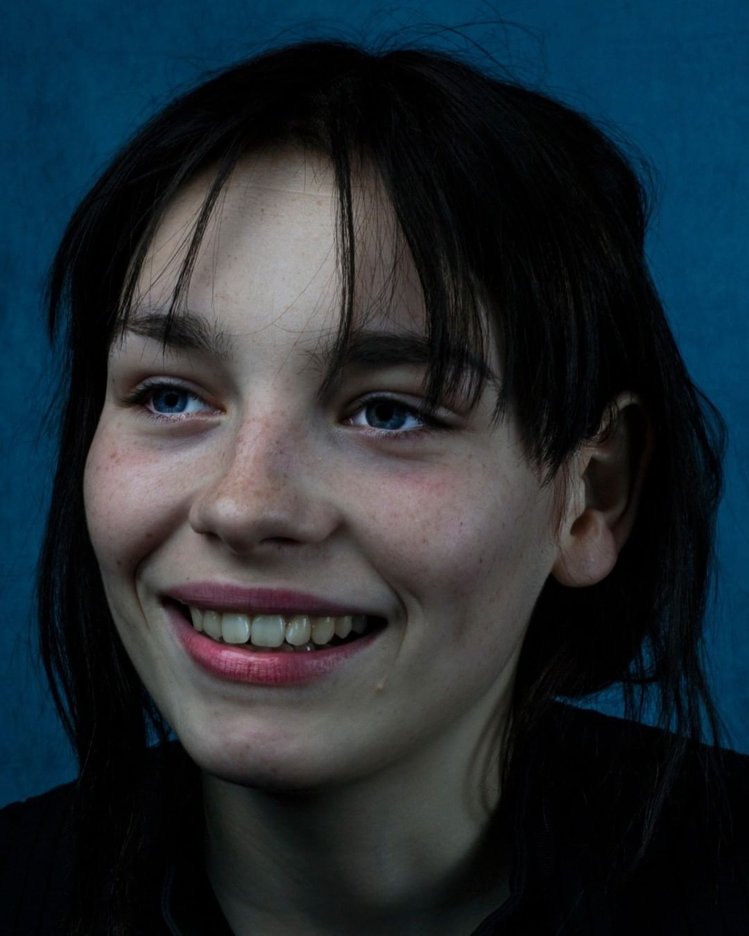 Close-up portrait of a smiling young woman with dark hair and blue eyes against a dark blue background. She is wearing a dark shirt.