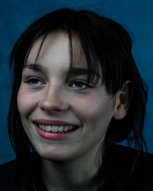 Close-up portrait of a smiling young woman with dark hair and blue eyes against a dark blue background. She is wearing a dark shirt.