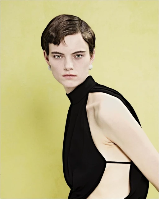 Portrait of a woman with dark, short hair wearing a black, high-necked dress against a yellow background in a studio environment.