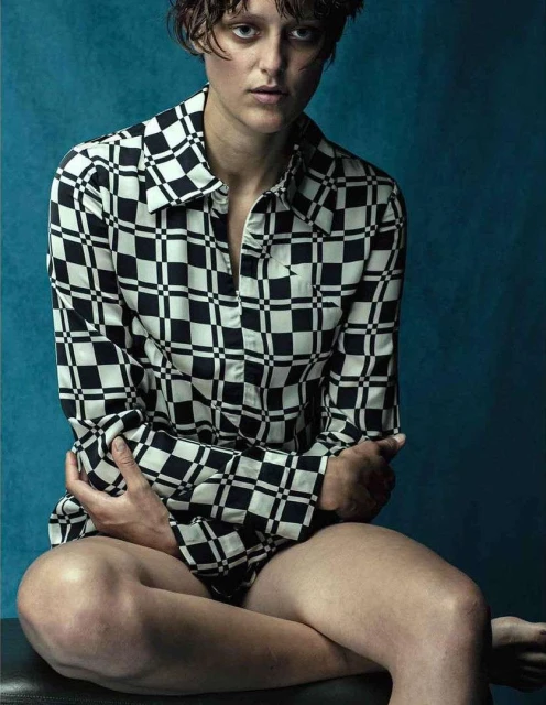 Young woman with wet dark hair poses sitting on a black stool in a studio wearing a black and white checkered shirt and bare legs against a dark teal backdrop.