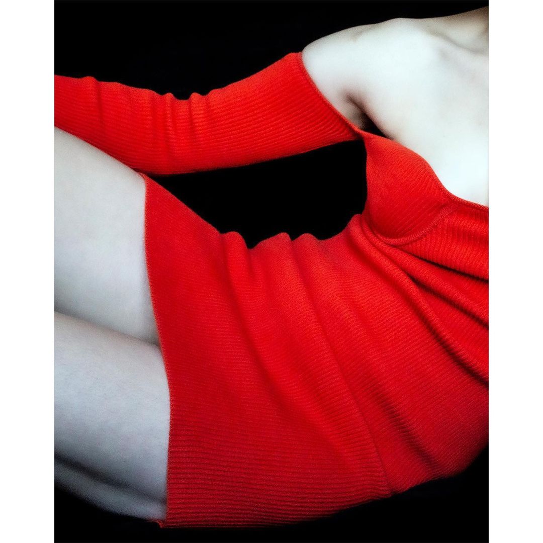 Close-up of a fair-skinned adult female wearing a short, red ribbed dress, set against a stark black background in a studio setting.