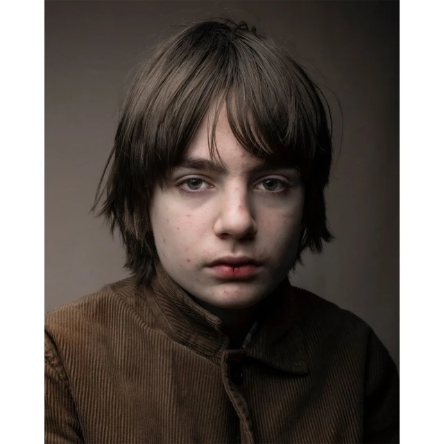 Portrait of a young boy with brown hair and eyes wearing a corduroy jacket in front of a neutral studio background, eye-level shot.