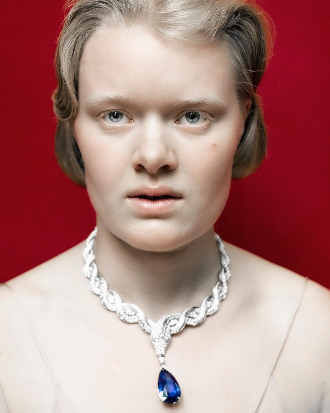 Portrait of a fair-skinned adult female with short blonde hair wearing a large silver necklace with a blue gemstone pendant against a red backdrop.