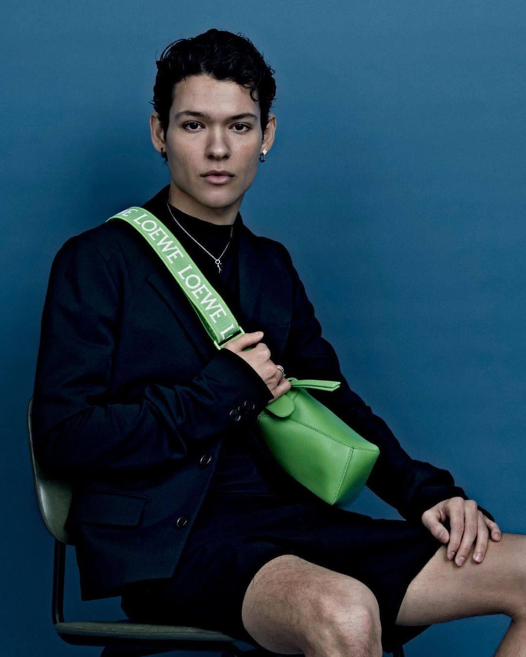 Person poses while seated in a black blazer with a lime green Loewe purse and strap against a blue backdrop in a medium studio shot.