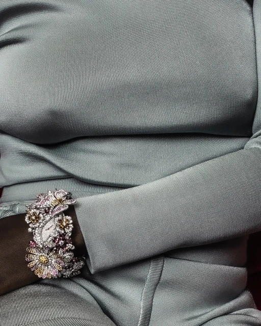 Closeup of a woman's arm wearing an ornate floral watch with jewels, against a backdrop of a silver dress with shiny fabric.