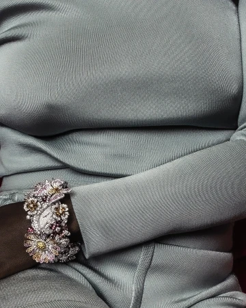 Closeup of a woman's arm wearing an ornate floral watch with jewels, against a backdrop of a silver dress with shiny fabric.