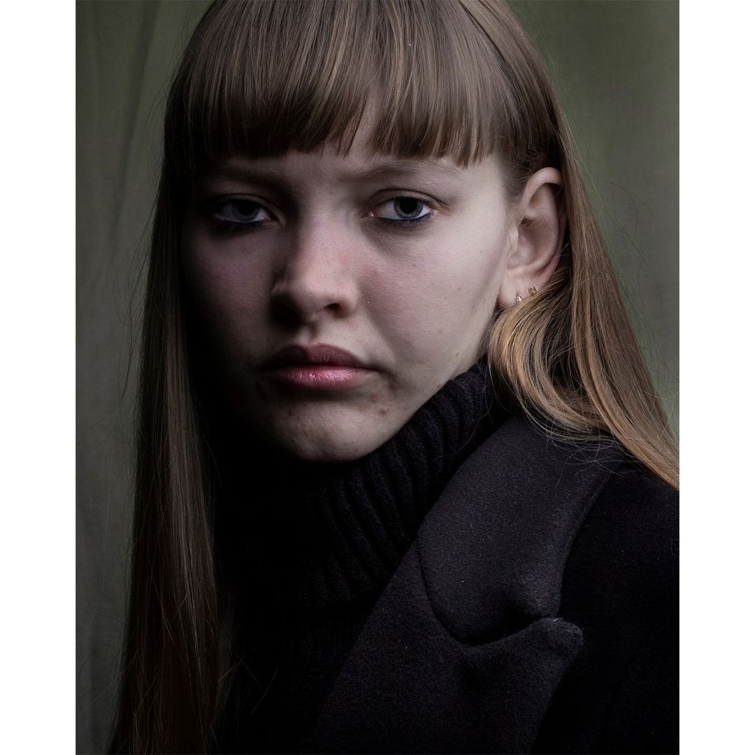 Portrait of a young woman with bangs looking at the camera. She is wearing a black turtleneck and a dark overcoat in a dimly lit indoor setting.