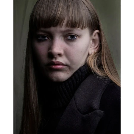 Portrait of a young woman with bangs looking at the camera. She is wearing a black turtleneck and a dark overcoat in a dimly lit indoor setting.