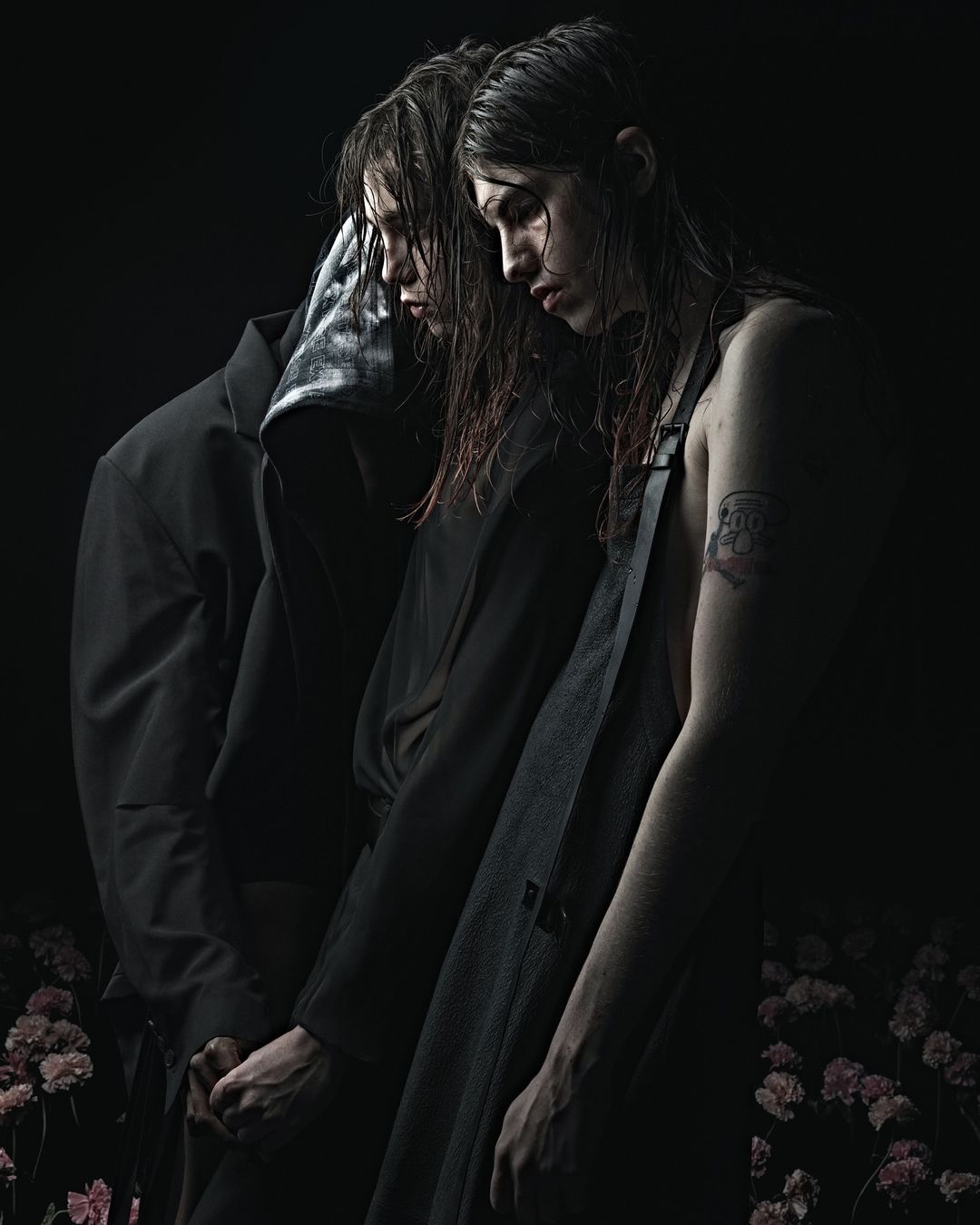 Two dark-haired people stand in close proximity amidst scattered pink flowers in a dimly lit, moody photograph, creating an intimate atmosphere.