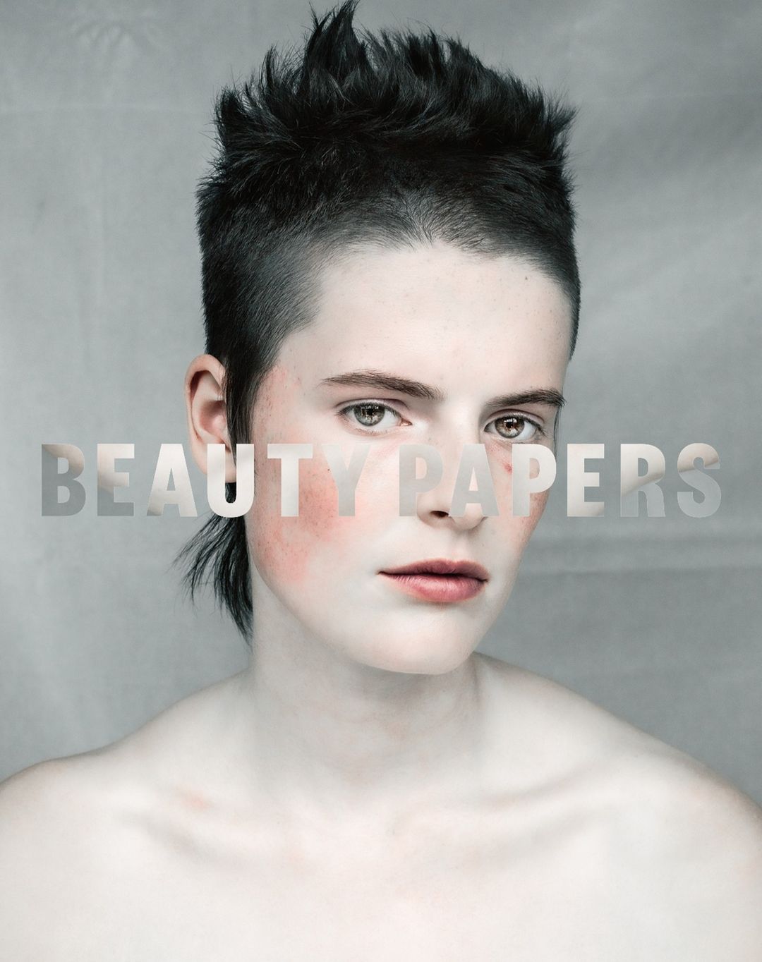 Close-up beauty shot of a young adult with a short, spiky haircut, wearing pale makeup and blush, against a neutral background.