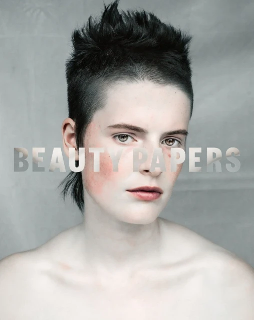 Close-up beauty shot of a young adult with a short, spiky haircut, wearing pale makeup and blush, against a neutral background.